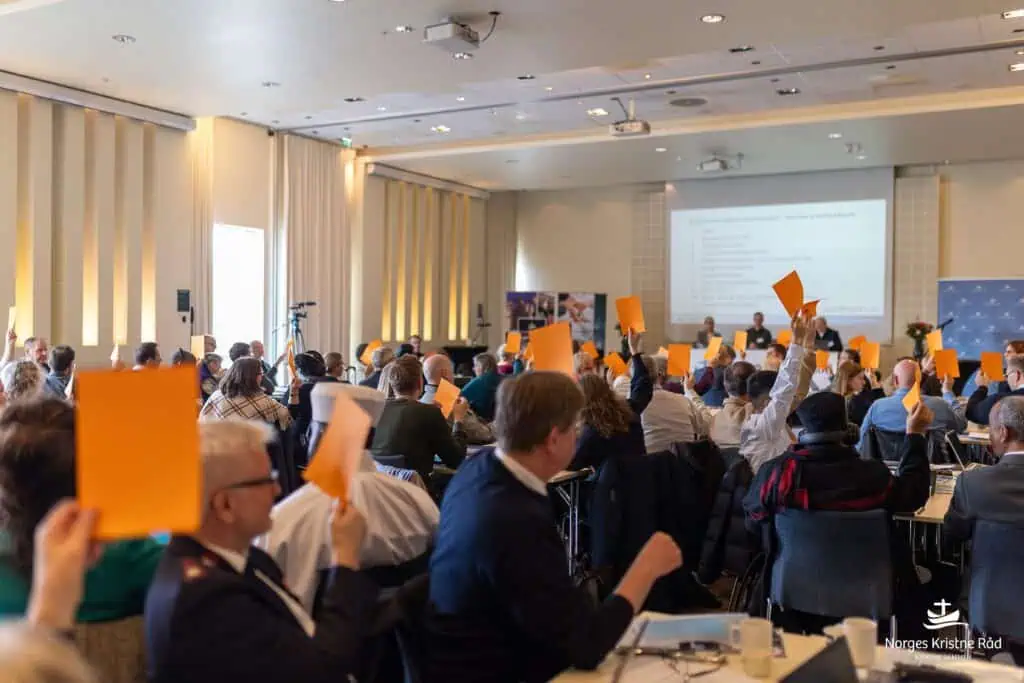 Bilde av en generalforsamling, der et mangfold av hender fra et mangfold av kirkesamfunn og etnisiteter holder lapper i været.