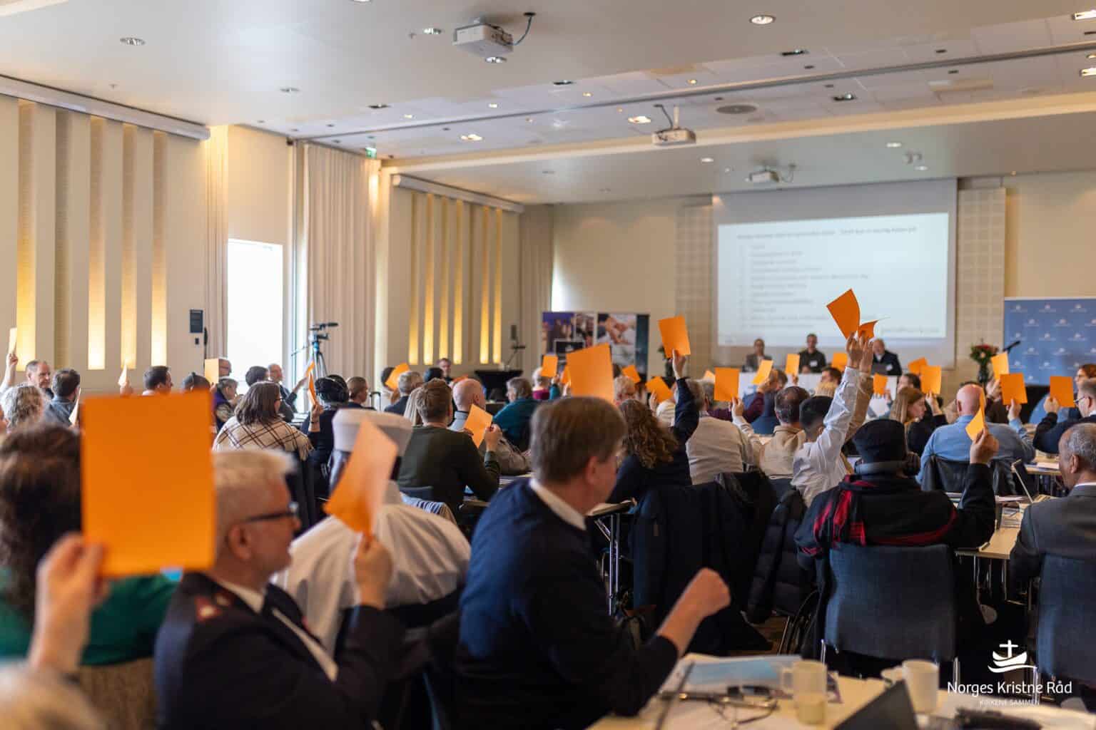 Bilde av en generalforsamling, der et mangfold av hender fra et mangfold av kirkesamfunn og etnisiteter holder lapper i været.